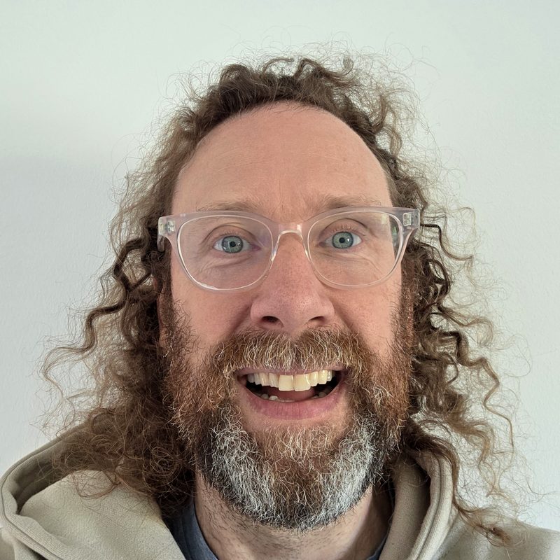 Ally Burns, smiling, with curly hair, glasses and a beard